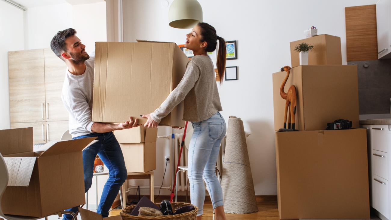 Man and woman carrying box through room amid other boxes and furniture