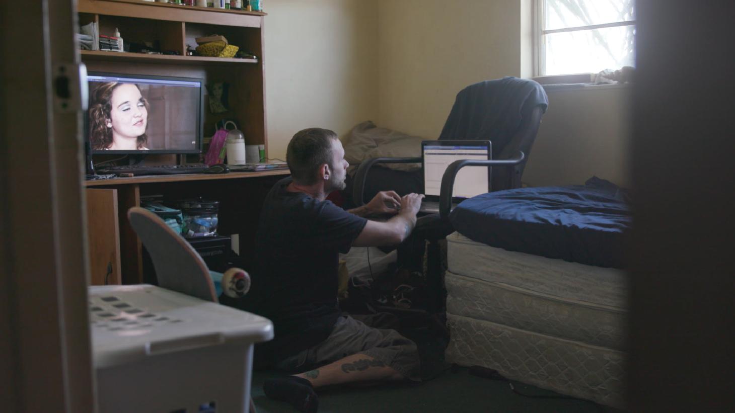 A man works on his computer while a video plays on another monitor elsewhere in his room. 