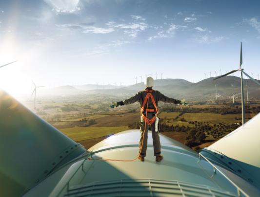 A person stands on a wind turbine, overlooking a wind farm. 