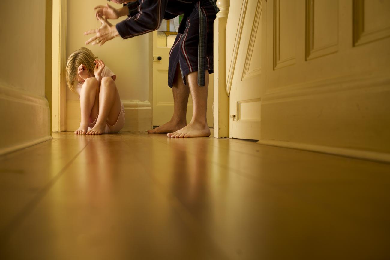 A child cowers in a corner while an adult's hands are positioned over her.