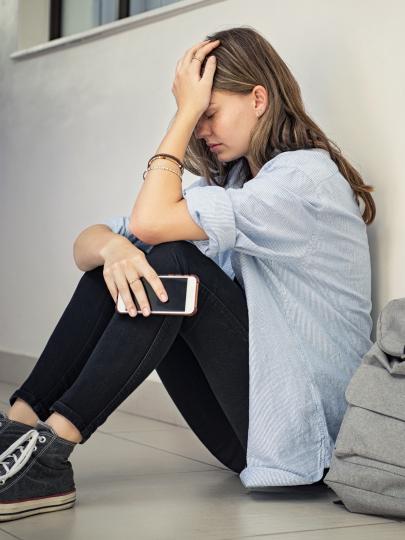 Young woman sitting against wall on hallway floor with hand on head and holding smartphone